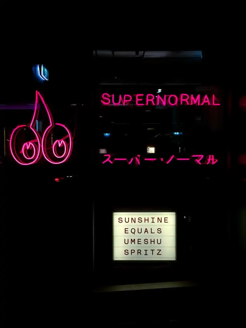 Black background, in neon pink is a sign that says Supernormal. Smaller sign says Sunshine equals umeshu spritz