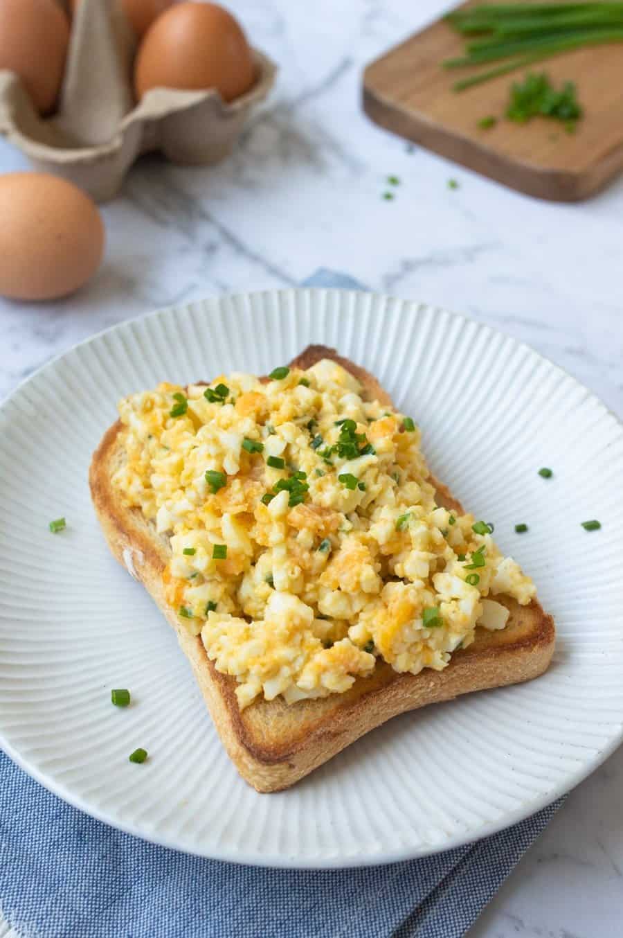 white plate in foreground with piece of toast topped with smashed eggs and garnished with chives.