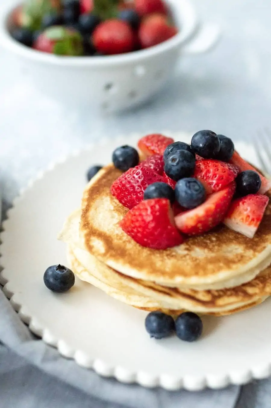 Scalloped plate with a stack of pancakes topped with strawberries and blueberries. In the background is a colander with more strawberries and blueberries