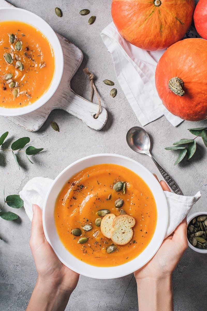 Image of orange pumpkins and a bowl of pumpkin soup on a table. Second bowl of pumpkin soup in hands being offered.