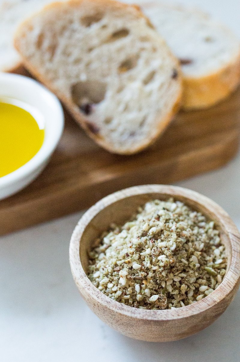 Small wooden bowl filled with nut free dukkah. In the background is some sliced sourdough bread and a bowl of olive oil