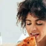 Woman of colour with curly hair about to bite into a piece of pizza. Text reads