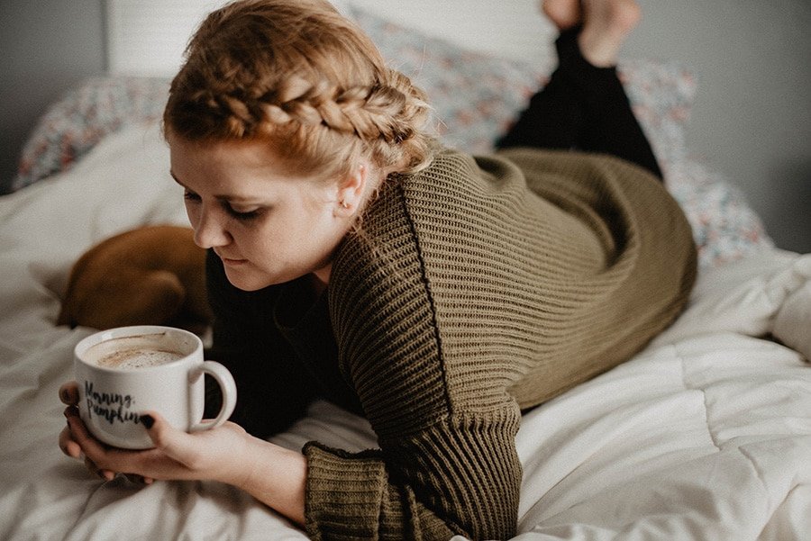 Girl lying on bed in olive green jumper with plaited hair holding a mug of coffee