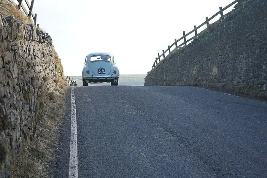 Light blue VW beetle driving on a road. The ocean is in the horizon