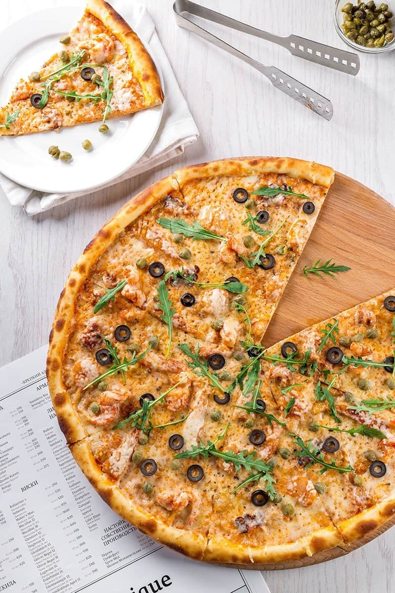 Image of a table top with a large pizza with olives, rocket, cheese, chicken and capers. There is a piece missing and is on a plate in the background