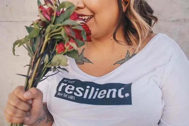 Female in larger body with red lipstick smelling a bunch of roses. She wears a white t-shirt that says "My size? Resilient. Drop the label"