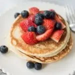 white scalloped edged plate with a stack of basic pancakes. Scattered over the pancakes are blueberries and sliced strawberries.