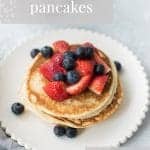 white scalloped edged plate with a stack of basic pancakes. Scattered over the pancakes are blueberries and sliced strawberries. Text overlay that reads basic pancakes. feel good eating