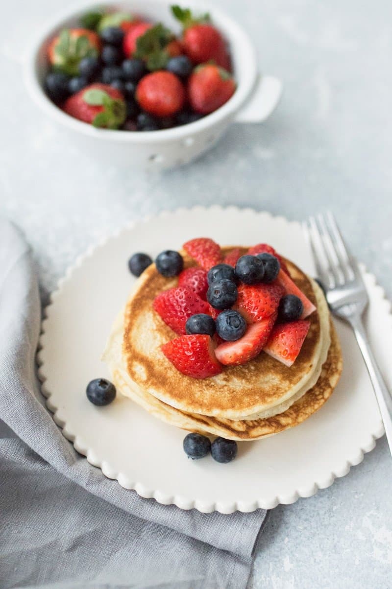 white scalloped edged plate with a stack of basic pancakes. Scattered over the pancakes are blueberries and sliced strawberries.