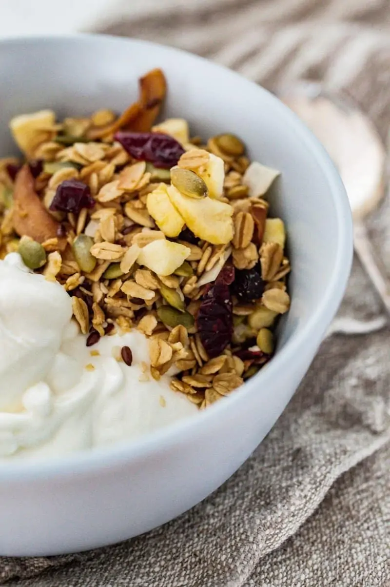 A blue bowl sitting on a grey striped tea towel is half filled with nut free granola and half filled with thick Greek yoghurt.