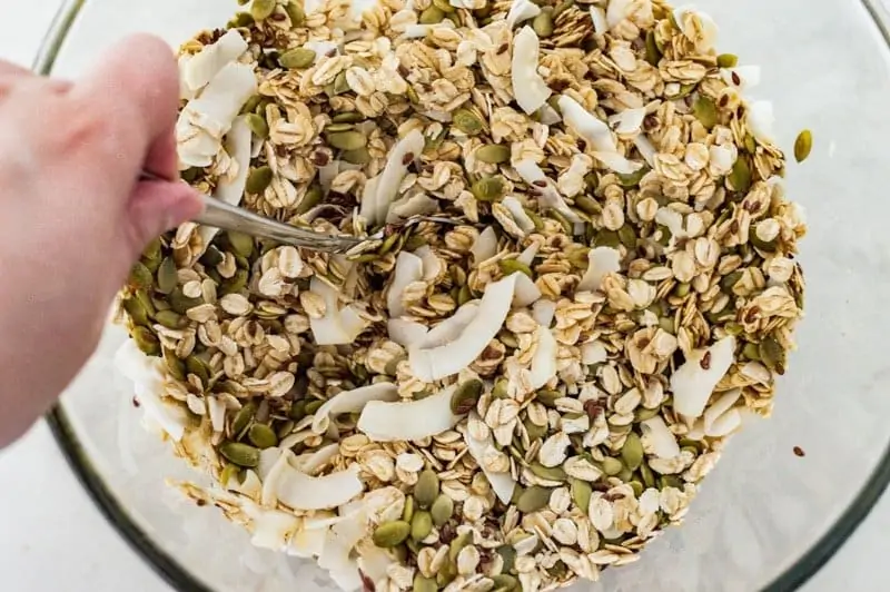 A person is mixing the nut free granola ingredients in a glass bowl.