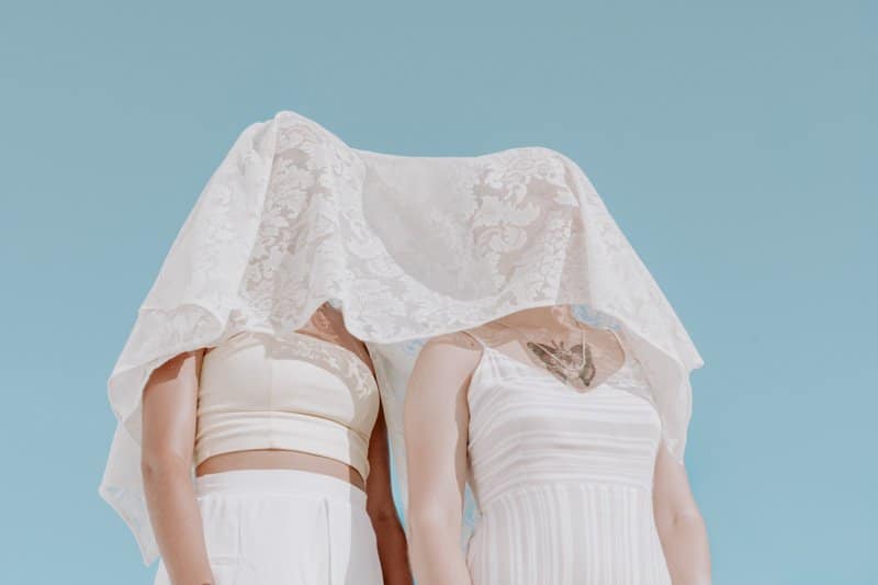 Two white females standing next to each other in white clothes. They have a lacy veil over their faces.