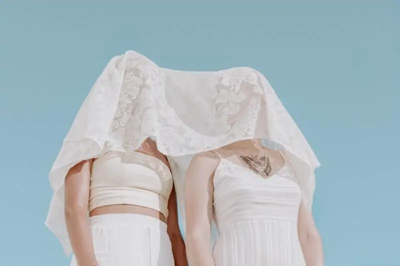 Two white females standing next to each other in white clothes. They have a lacy veil over their faces.