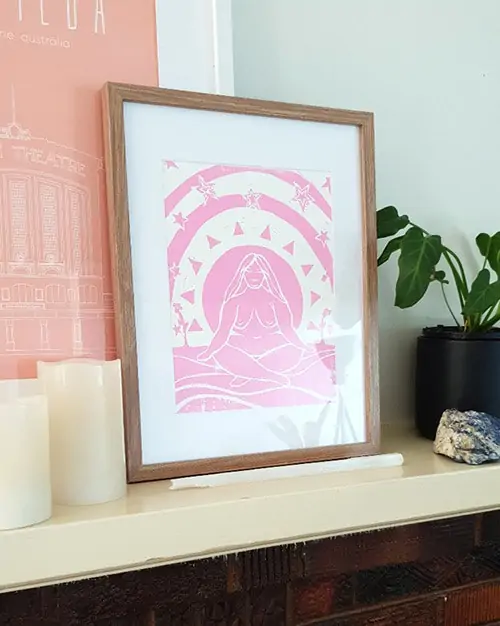 A hand printed image of a fat female-looking human surrounded by abstract shapes. The print is in pink ink and is in a light coloured wooden frame on a mantle.