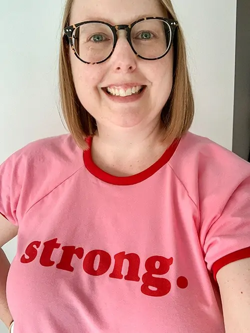 A smiling woman with shoulder length dark blonde hair and big round glasses. She wears a bright pink and red t-shirt that reads Strong.