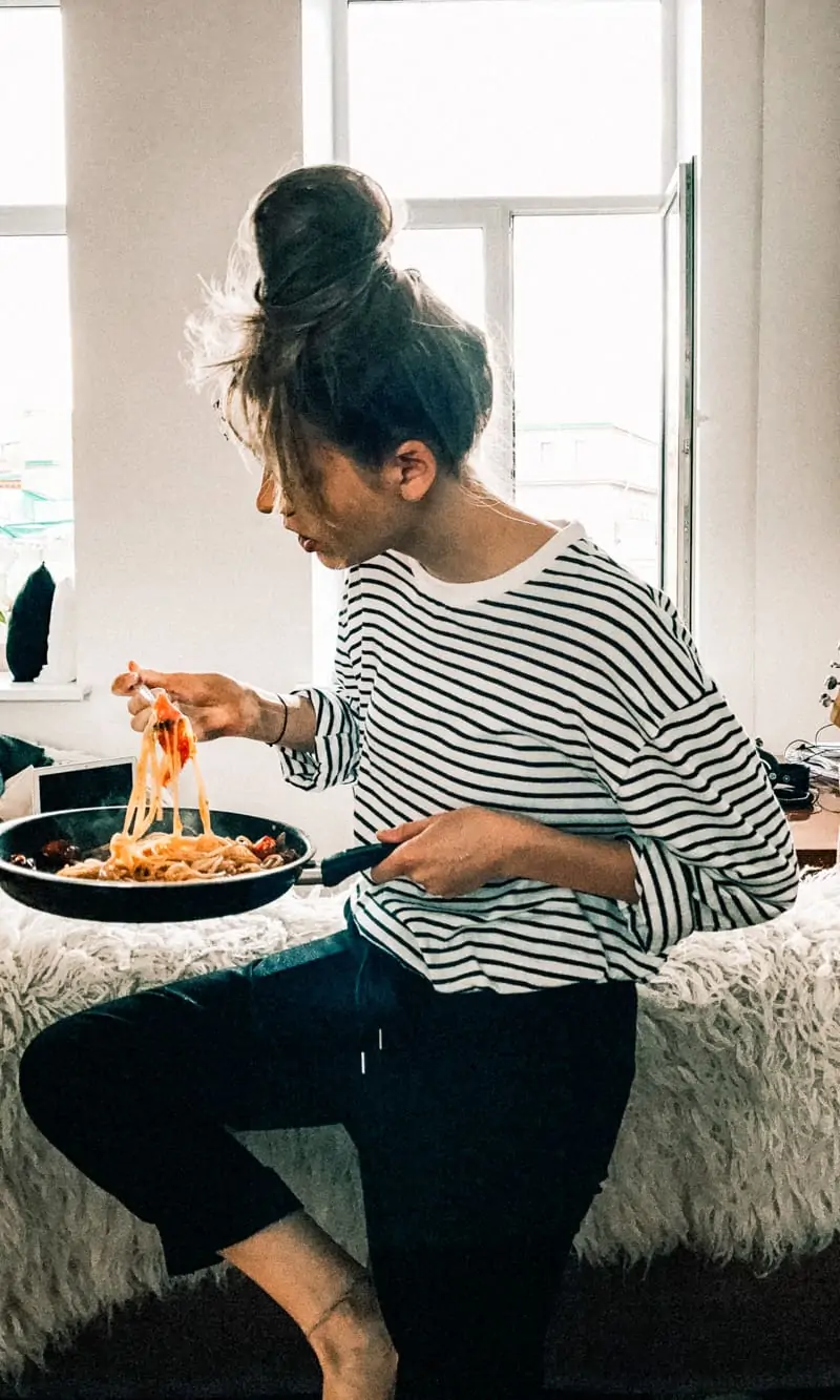 A woman in a striped shirt leaning on the back of a couch eating a whole pan of pasta.