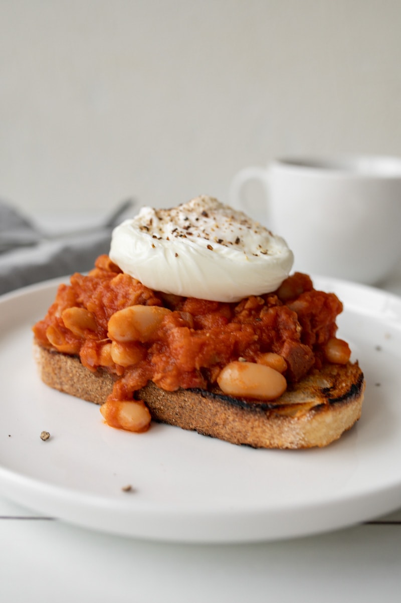 A plate with a piece of toast topped with smoky baked beans and a poached egg. There is a cup of coffee, espresso pot, eggs and tea towel in the background