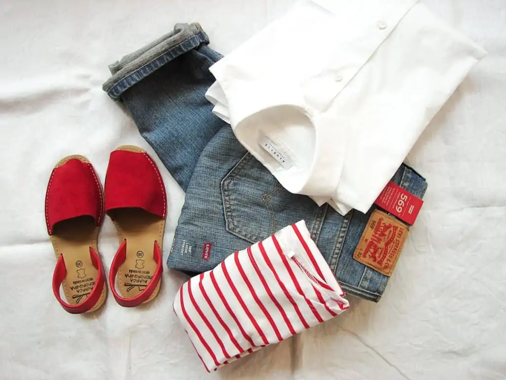 A pair of jeans, red and white striped top, white shirt and red espadrilles on a bed.
